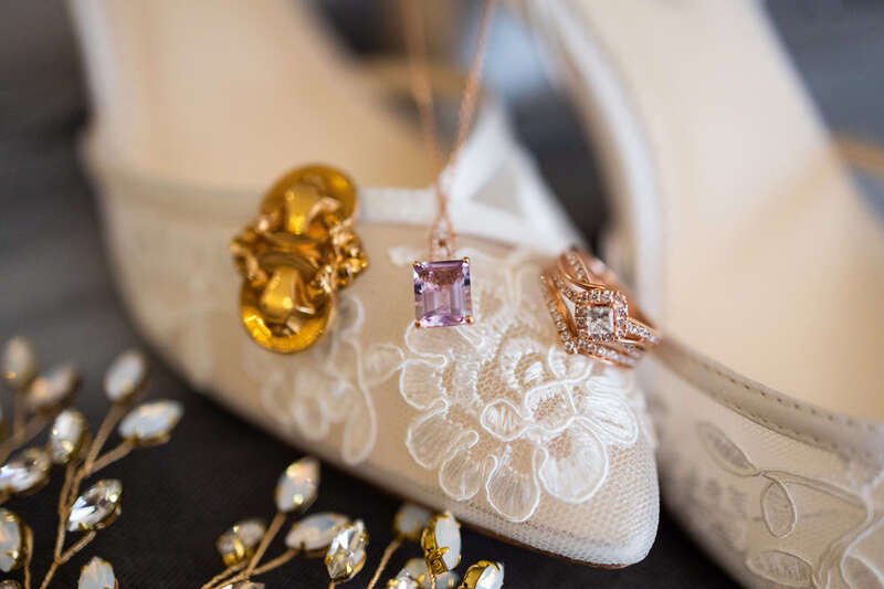 A close-up of lace wedding shoes with an engagement ring and amethyst pendant