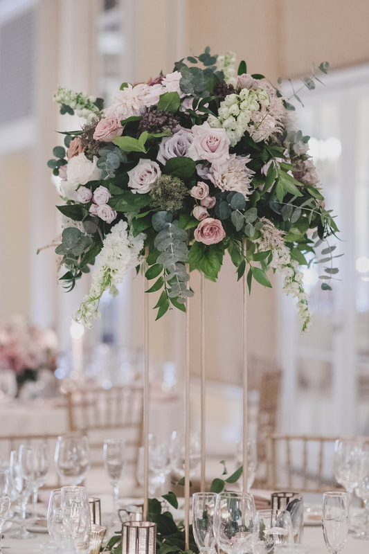 Tall floral centerpiece with light pink, light purple and white roses along with hanging greenery on a clear 4 columned pedestal 