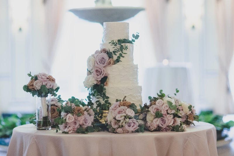 4 tiered white wedding cake with flowers draping around and the cake table lined with other floral bouquets and arrangements